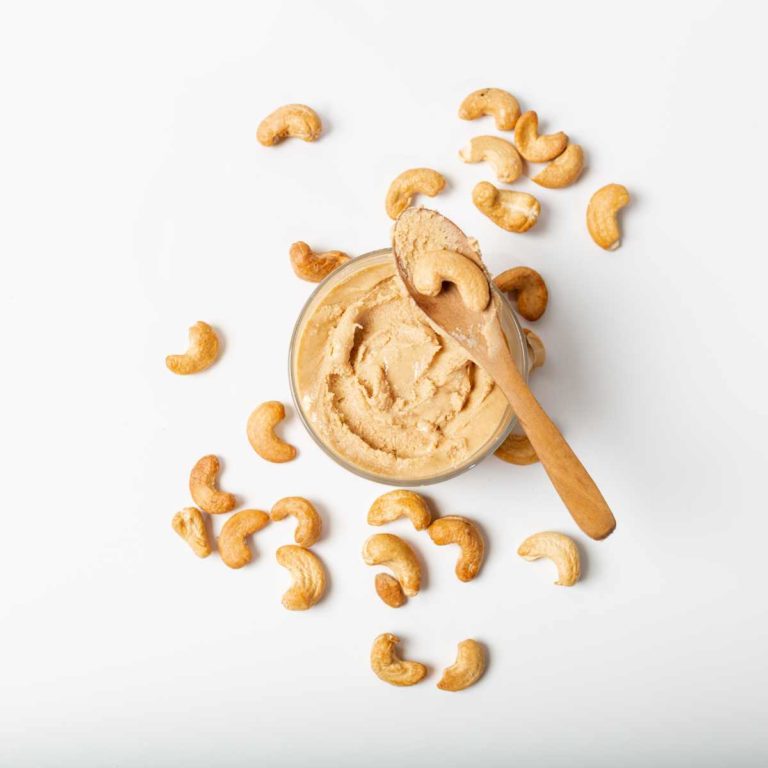 Glass jar of cashew paste with wooden spoon and whole cashew nuts scattered