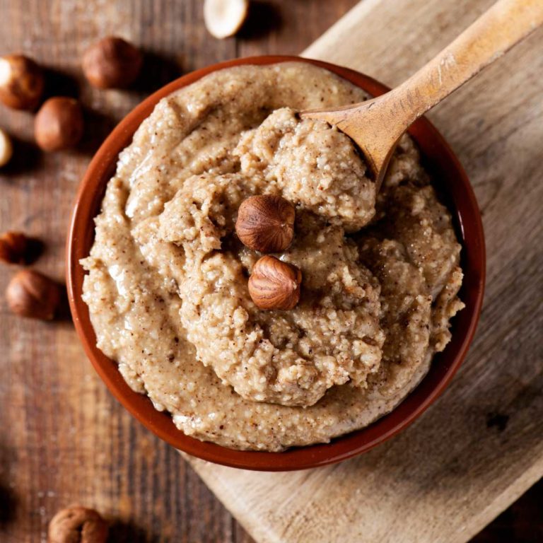 Light brown hazelnut paste in a bowl with spoon scooping it up. Hazelnuts dotted around