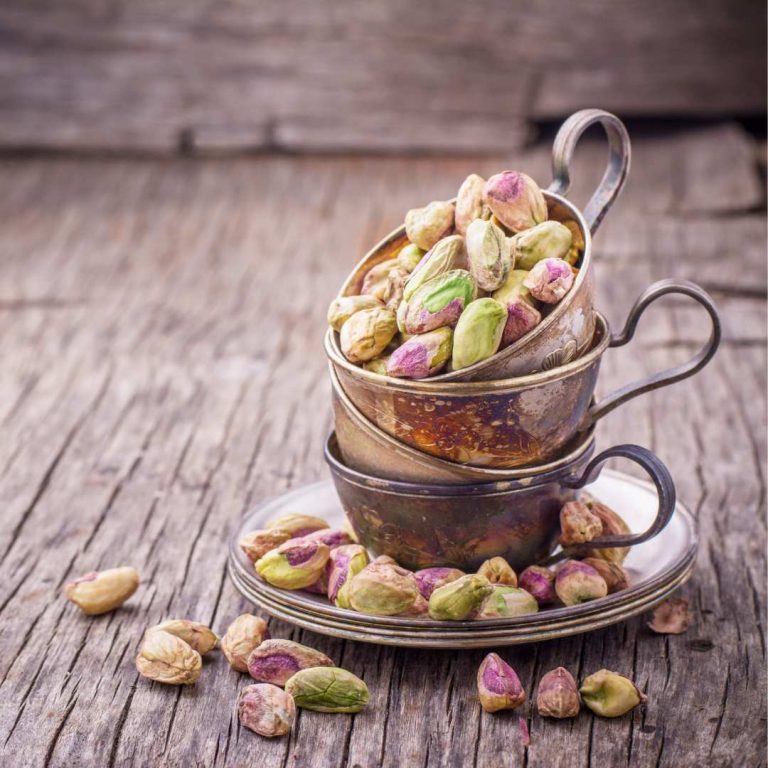 Shelled Pistachios in copper tea cups stacked on top of each other with saucers underneath, on a wooden surface.
